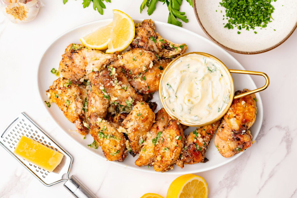 A platter of seasoned chicken wings garnished with herbs and parmesan, served with a bowl of creamy dipping sauce and lemon wedges.