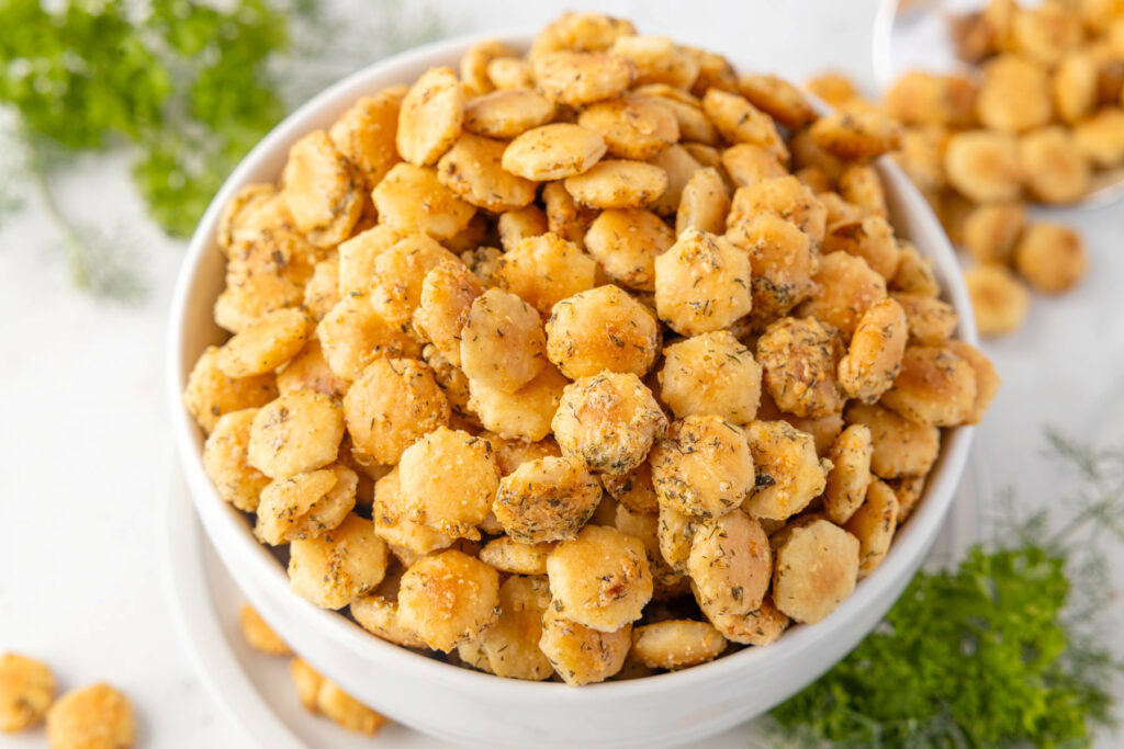 A white bowl filled with seasoned oyster crackers sits on a white surface, with herbs and more crackers scattered around.