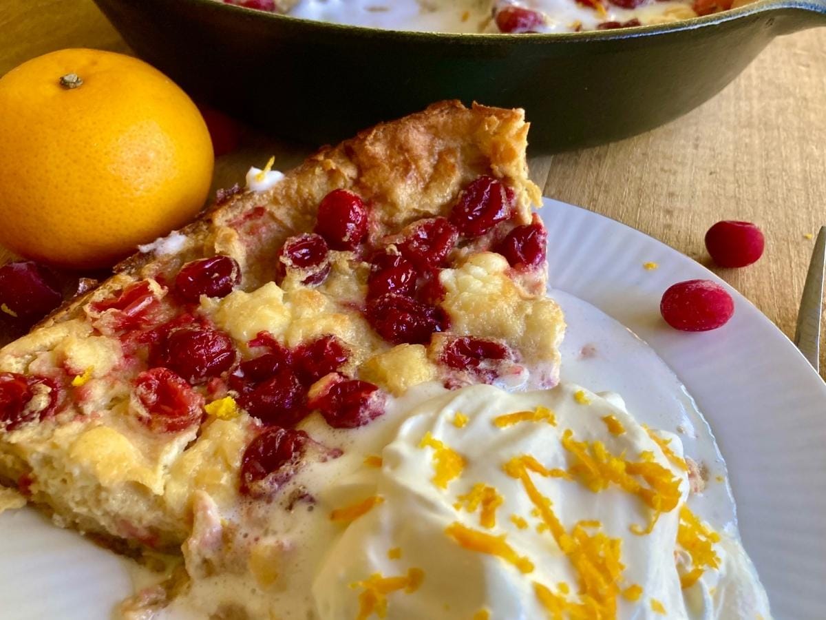A slice of cranberry clafoutis on a plate with whipped cream topped with orange zest, a fresh orange, and more clafoutis in a skillet in the background.