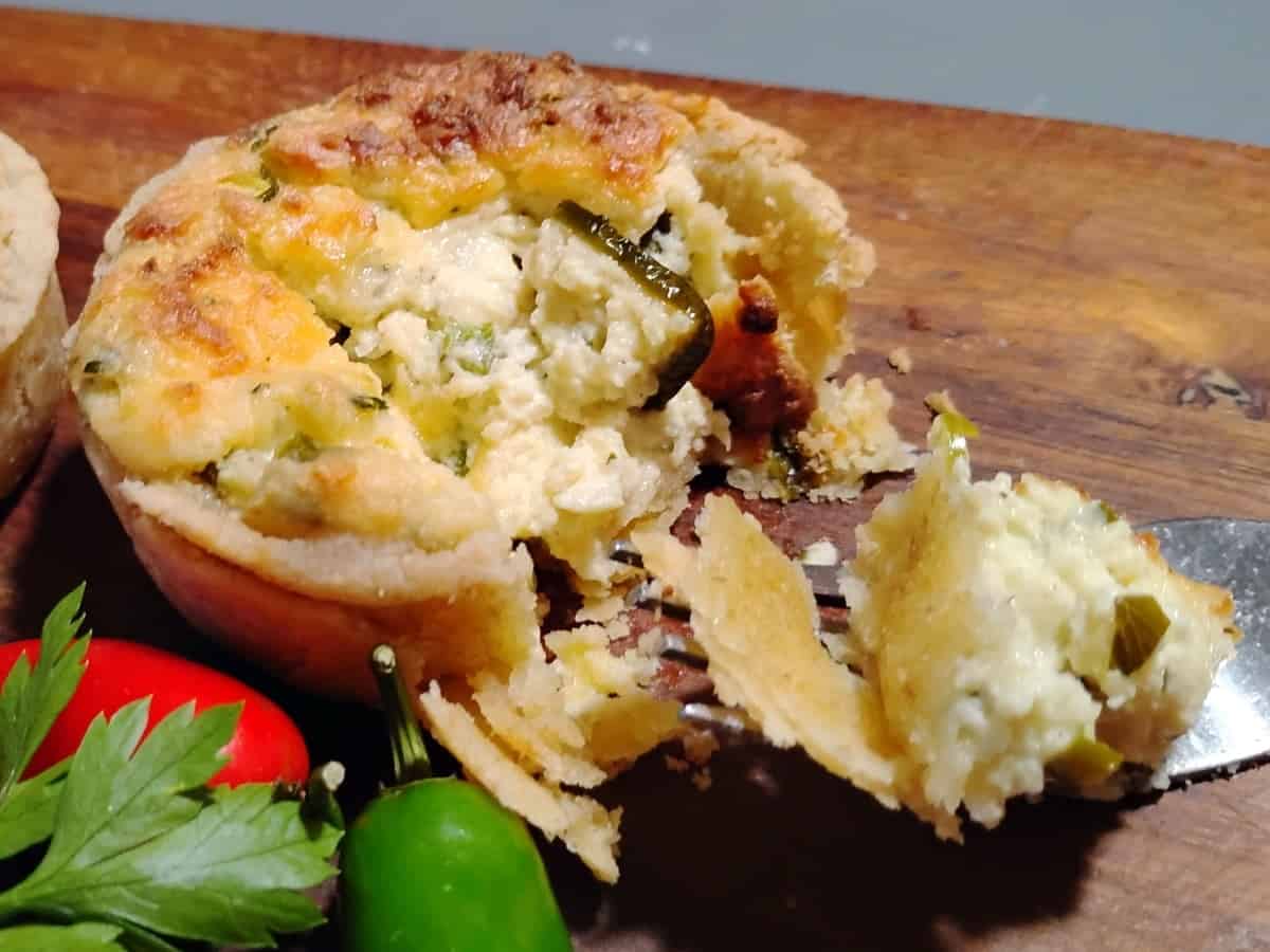 A close-up of a partially eaten savory pie with a flaky crust and cheesy filling, served on a wooden board with parsley and a chili pepper nearby.