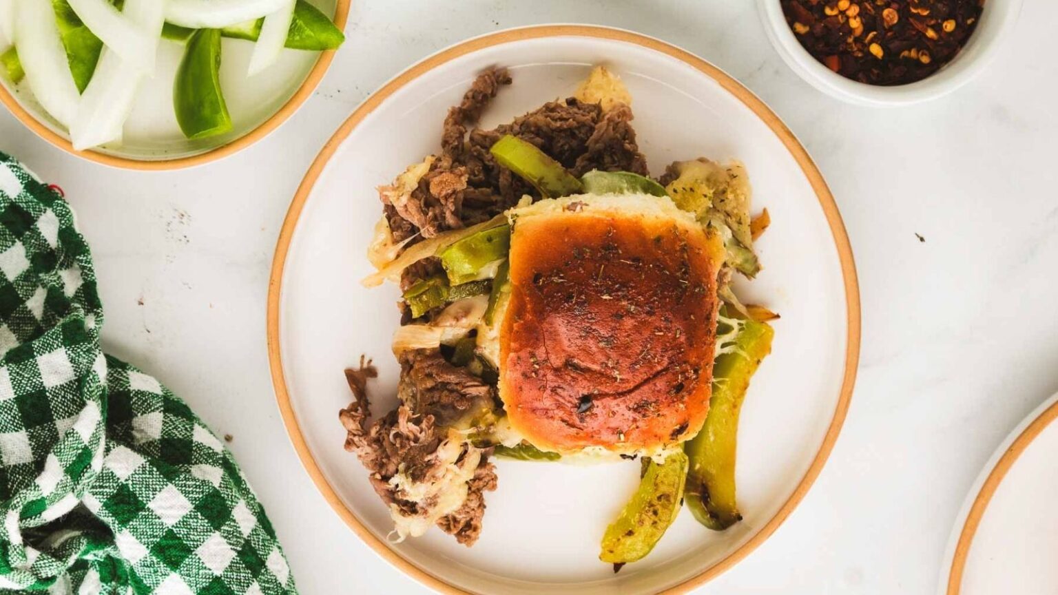 A plate with a baked sandwich topped with a golden bun, shredded beef, green bell peppers, and melted cheese, next to a green checkered napkin and bowls of sliced onions and chili flakes.