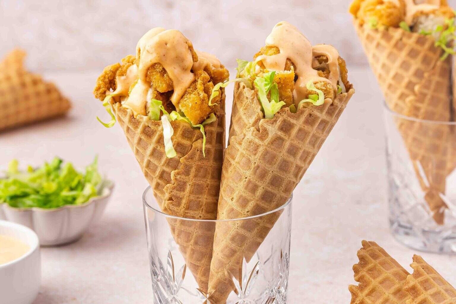 Two waffle cones filled with fried chicken, shredded lettuce, and creamy sauce are placed upright in clear glasses.