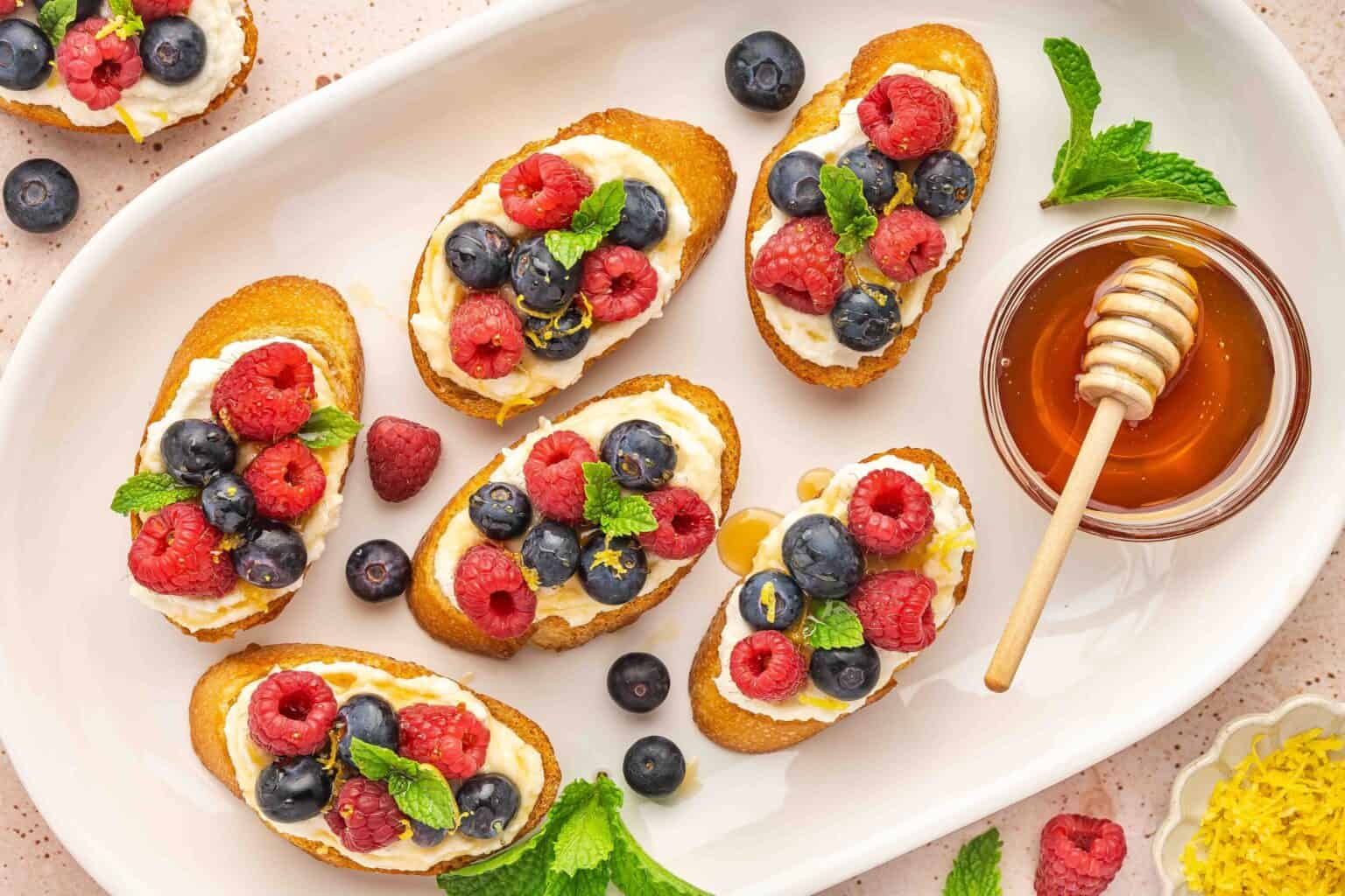 A plate of toasted bread slices topped with cream, raspberries, blueberries, mint leaves, and lemon zest, served with a honey dipper and fresh mint on the side.