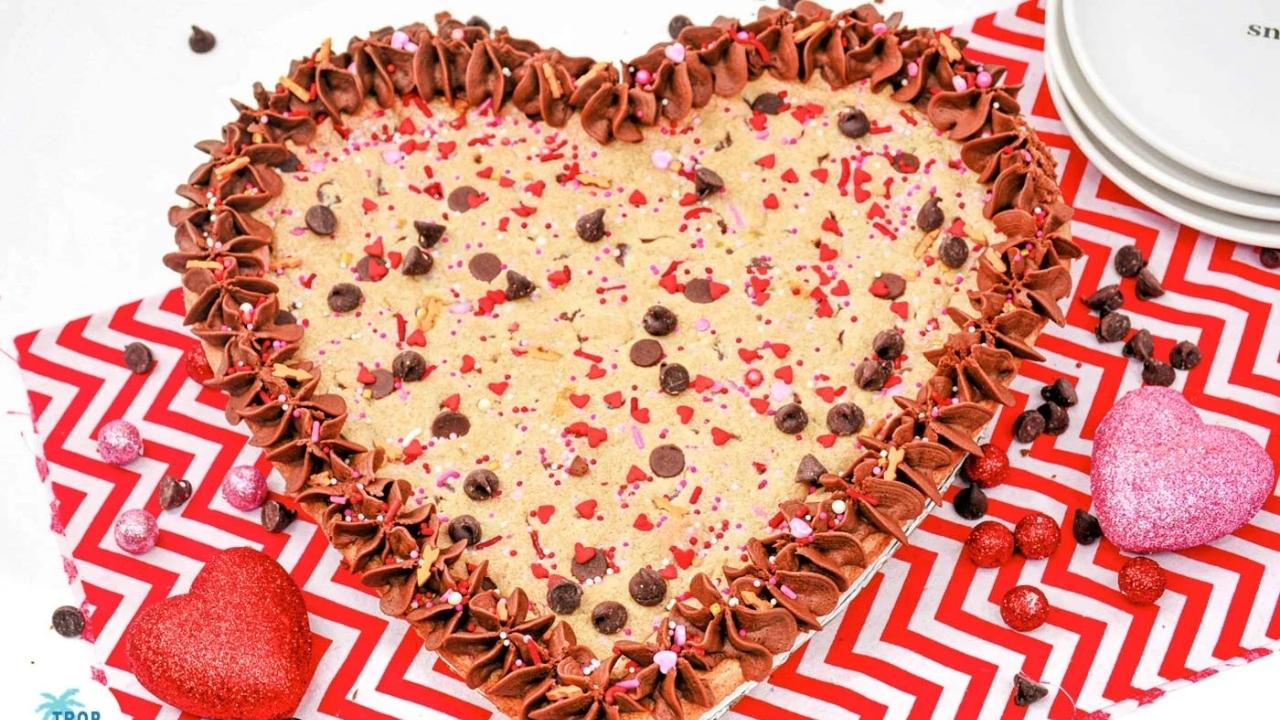 A heart-shaped cookie cake with chocolate frosting, chocolate chips, and red and pink sprinkles on a red chevron-patterned cloth, with decorative hearts nearby.