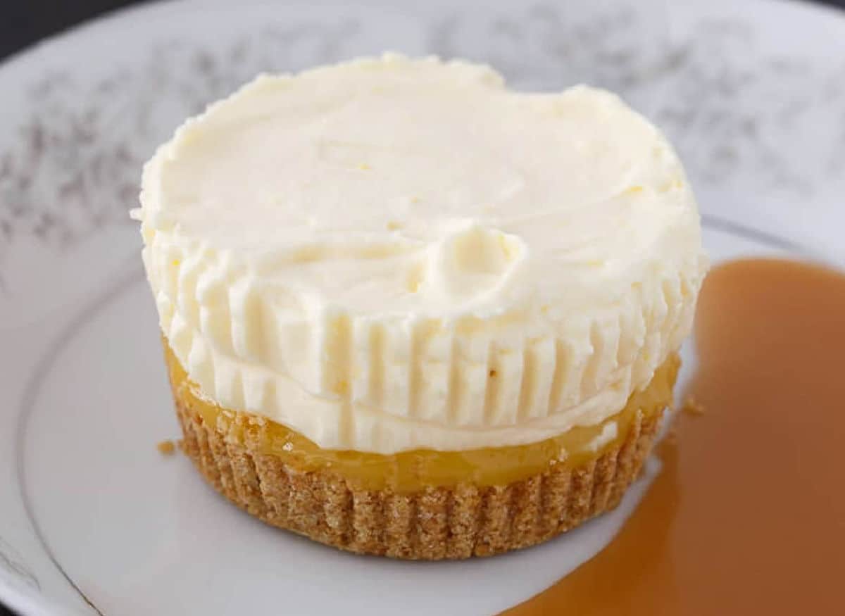A small cheesecake with a graham cracker crust, lemon filling, and creamy topping sits on a plate next to a pool of caramel sauce.