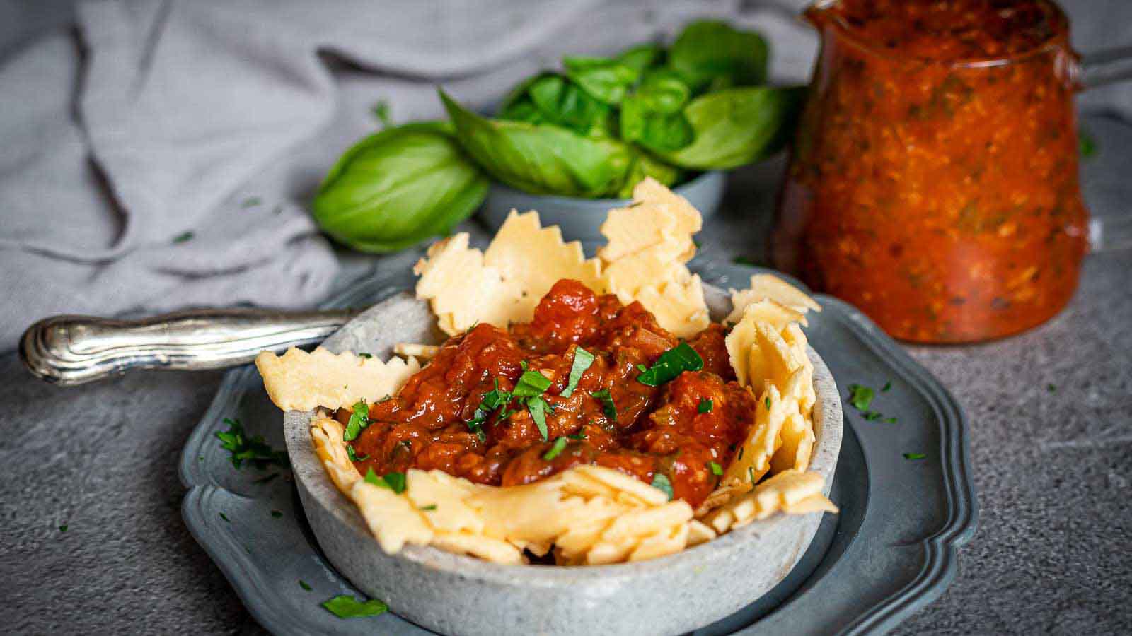A bowl of pasta with tomato sauce, garnished with parsley, sits on a plate with a fork. Fresh basil and a jar of extra sauce are in the background.