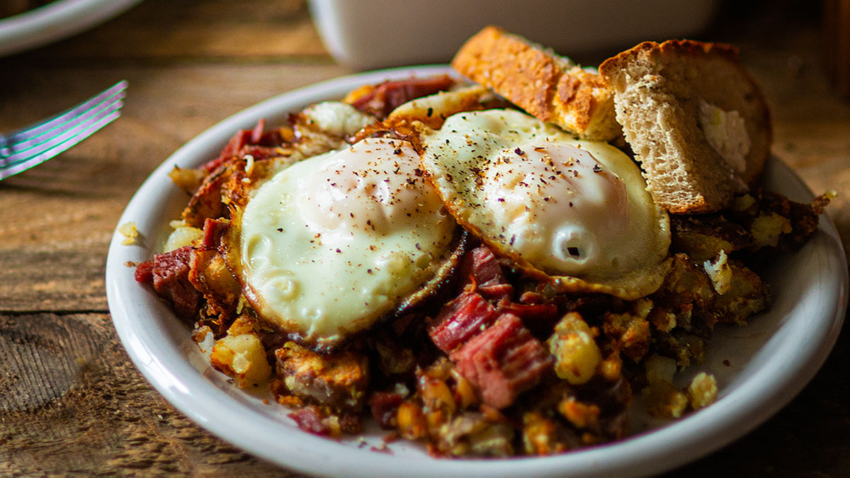 A plate of corned beef hash topped with two fried eggs, served with two slices of toasted bread on a wooden table.