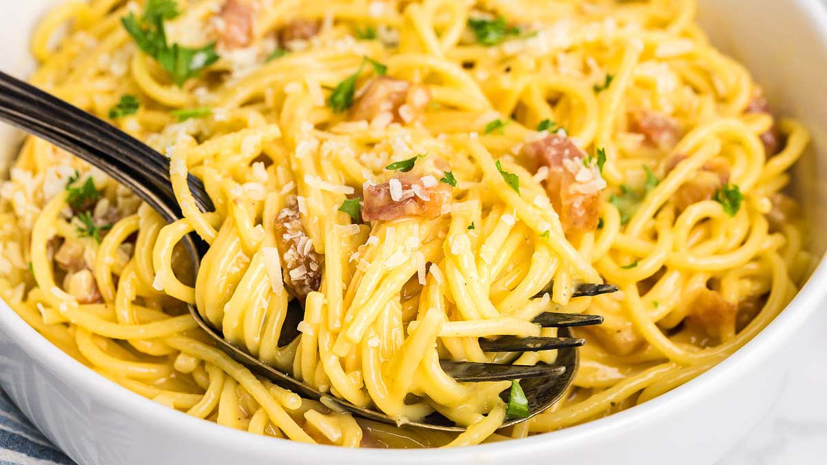 A close-up of spaghetti carbonara in a white bowl, garnished with parsley and grated cheese, with a fork twirling some pasta.