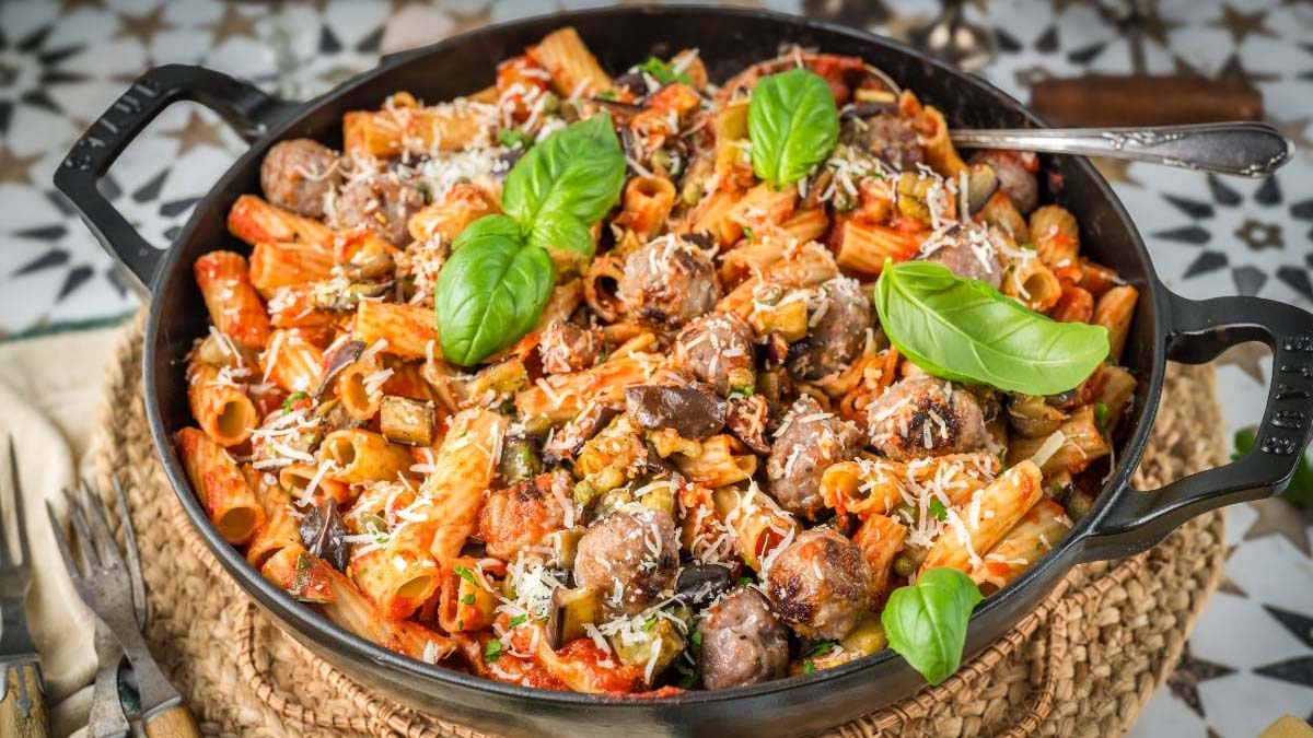 A black pan filled with rigatoni pasta, sausage, mushrooms, tomato sauce, grated cheese, and fresh basil leaves on a woven placemat.