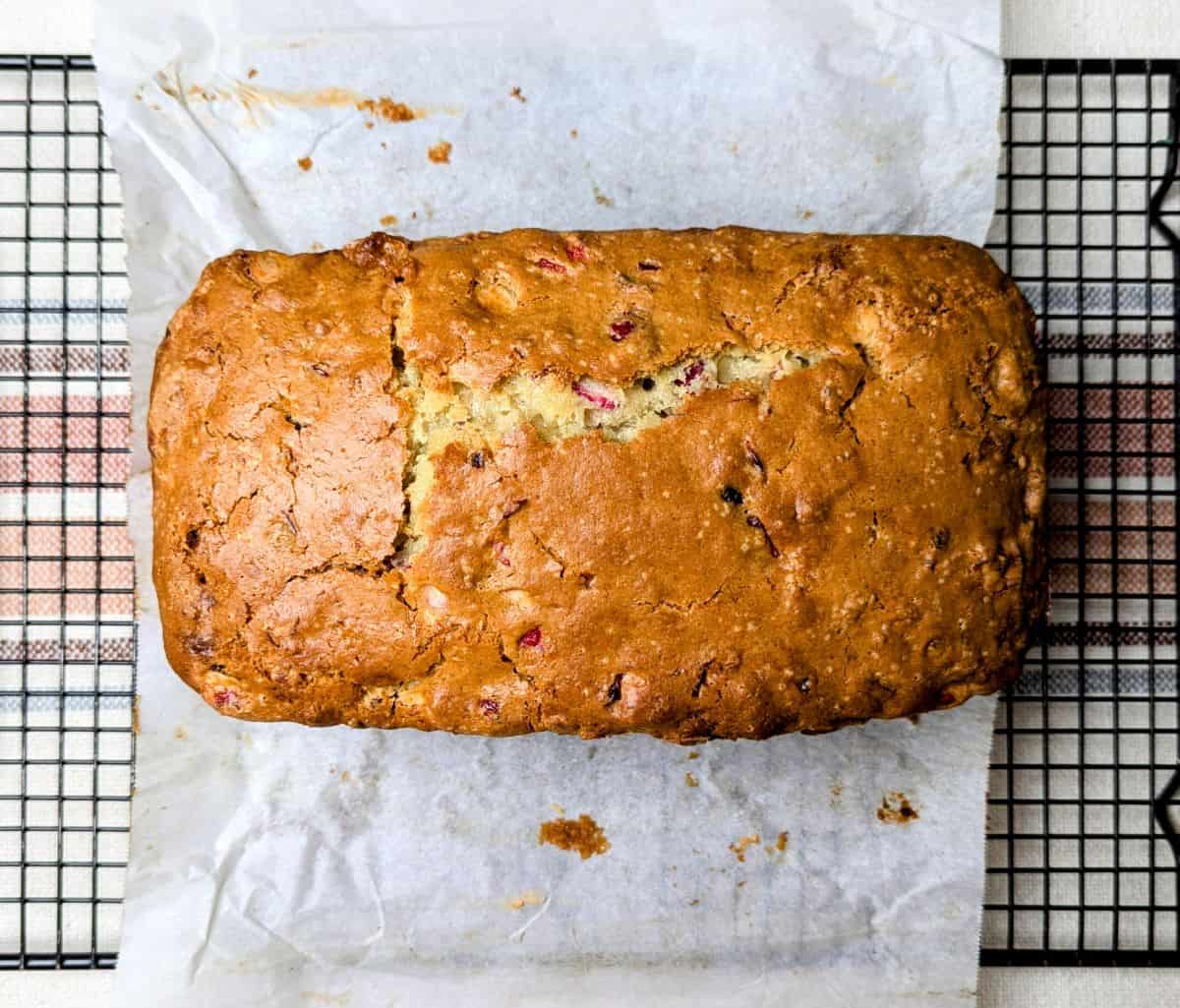 Loaf of golden brown bread with visible fruit pieces, resting on parchment paper atop a wire cooling rack.