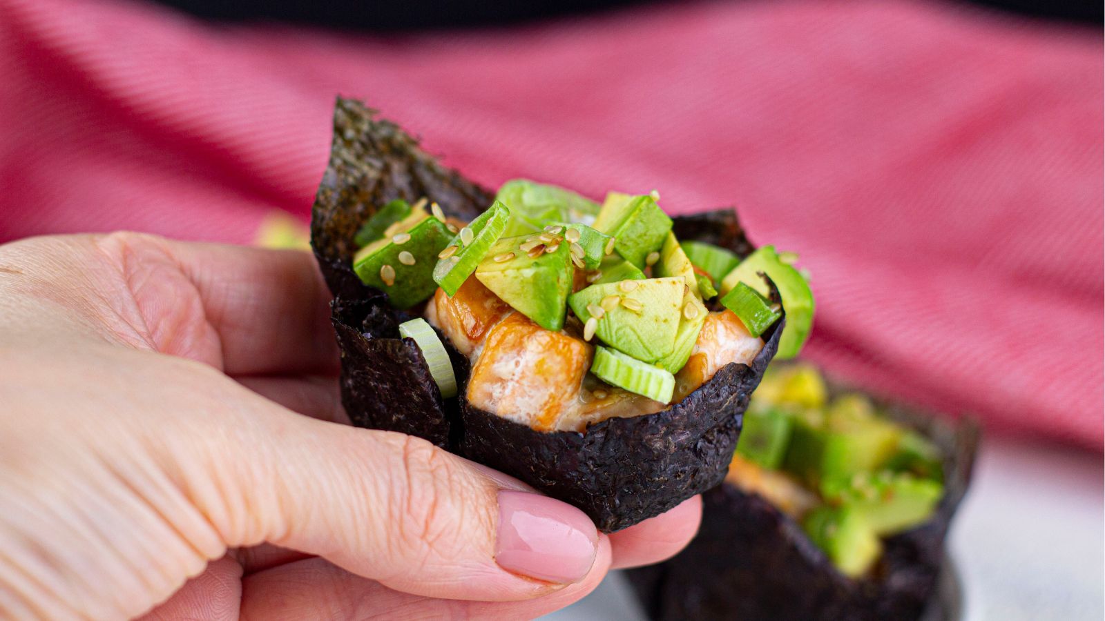 A hand holds a sushi hand roll with nori, rice, avocado, cucumber, and sesame seeds, with a red striped cloth in the background.
