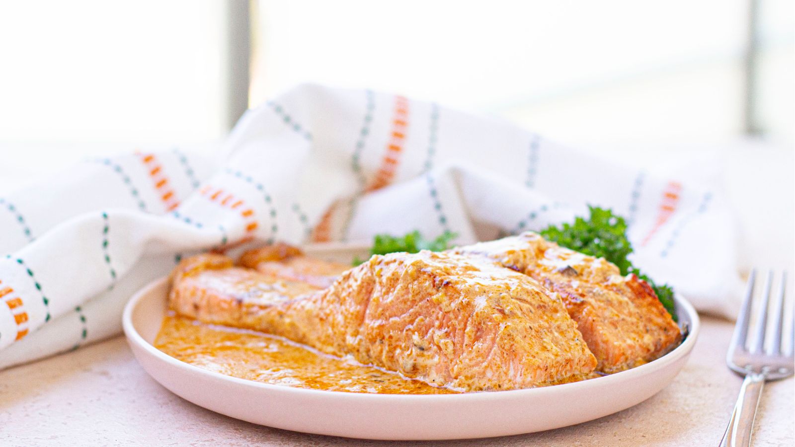 A cooked salmon fillet with creamy sauce is served on a white plate, garnished with parsley, with a striped cloth in the background.