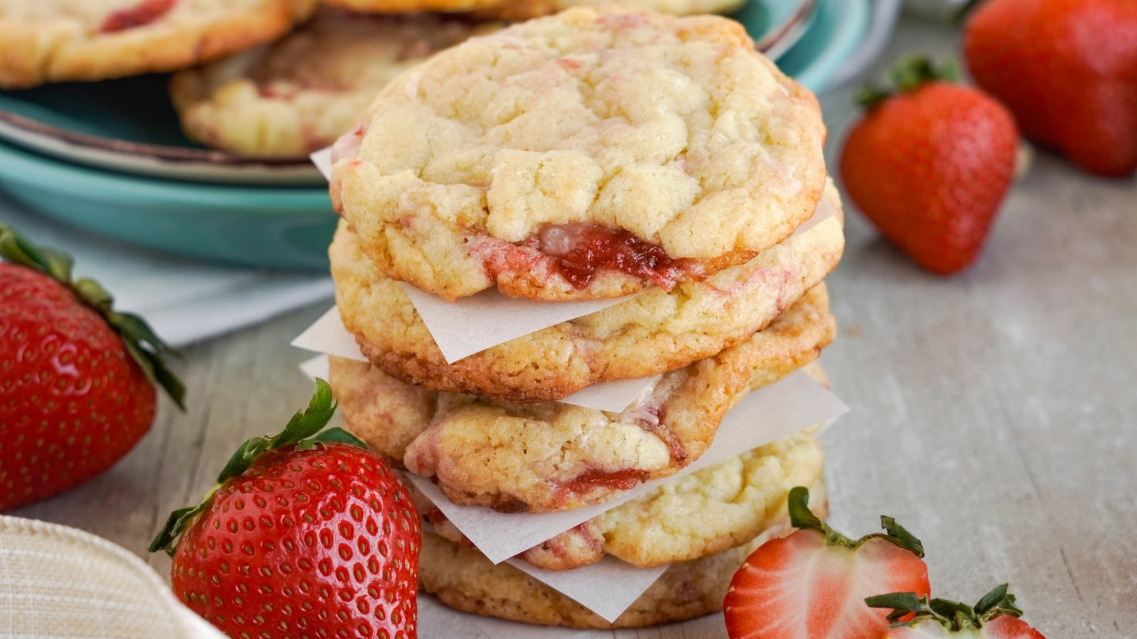 A stack of three strawberry cookies with pieces of parchment paper between them, surrounded by fresh strawberries on a wooden surface.