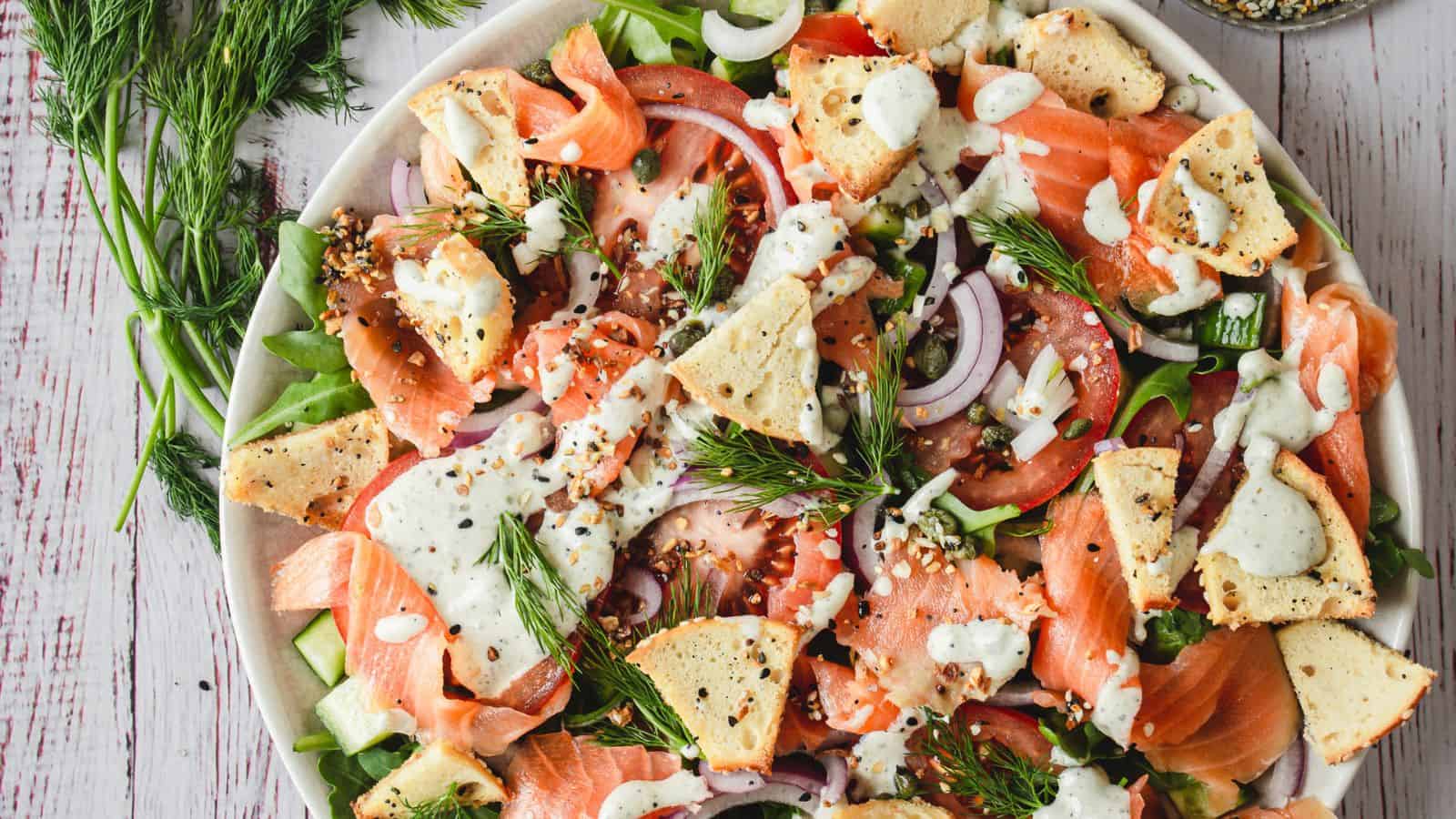 A plate of salad with smoked salmon, tomato slices, red onion, pita pieces, fresh dill, and a white dressing on top.