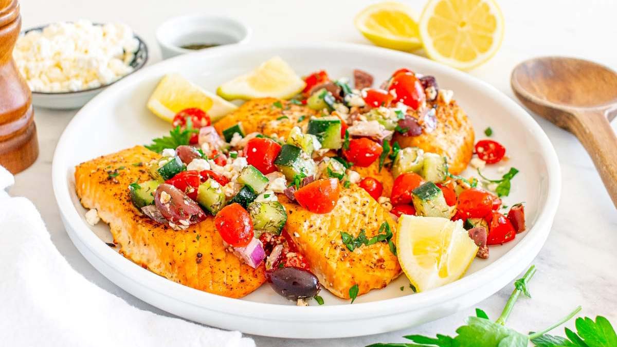 Grilled salmon fillets topped with a Mediterranean salad of tomatoes, cucumbers, olives, and feta cheese, garnished with lemon wedges, served on a white plate.