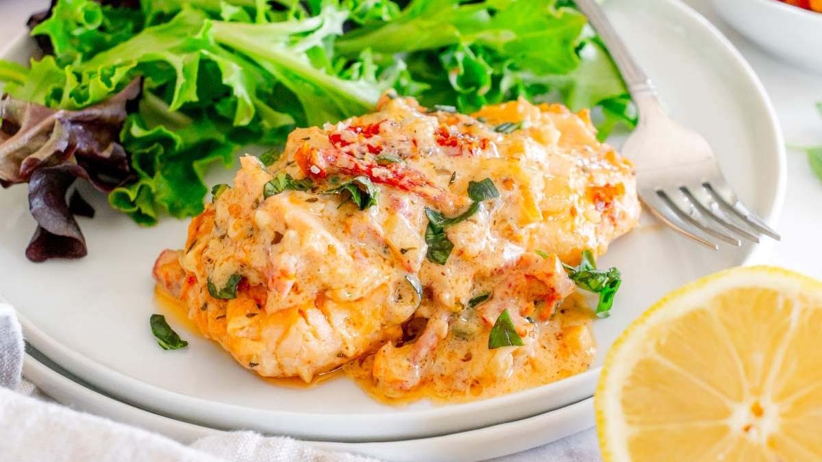 A serving of creamy, baked seafood dish garnished with herbs, plated next to fresh mixed greens, with a lemon slice and a fork on the side.