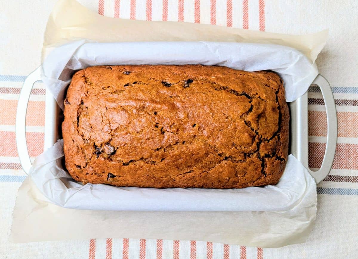 A loaf of banana bread in a parchment-lined baking pan, sitting on a striped cloth.