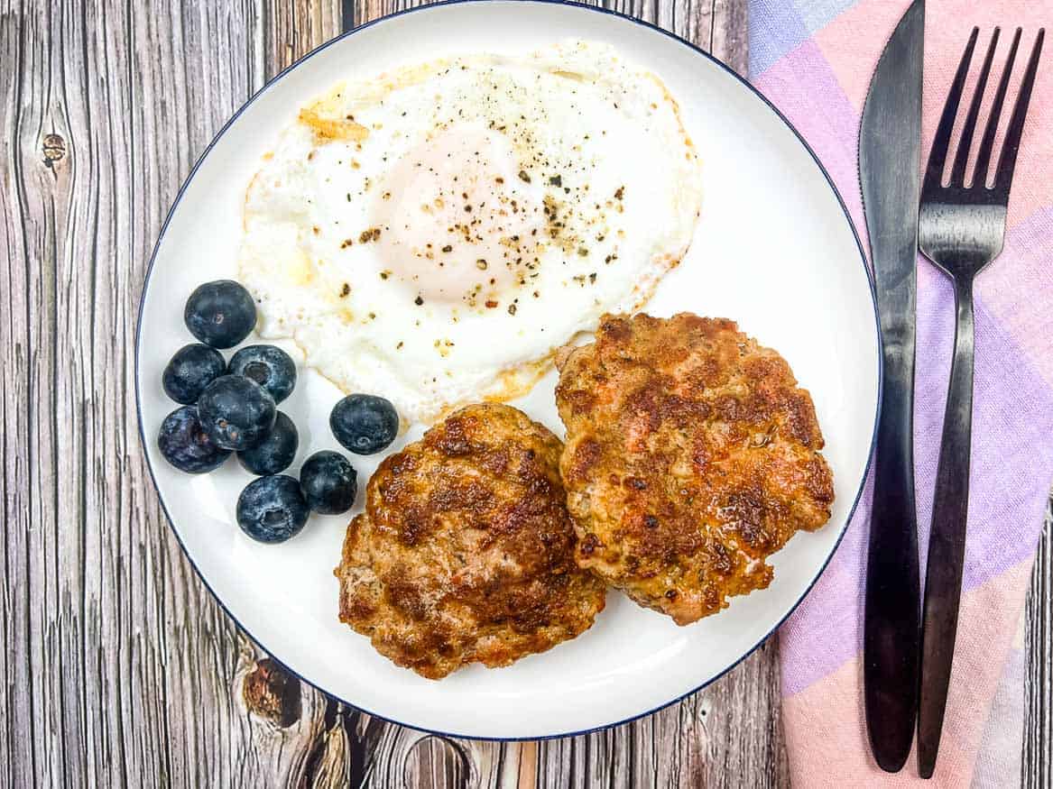 A plate with a fried egg, two sausage patties, and a small pile of blueberries, with a fork and knife beside a pastel napkin on a wooden table.