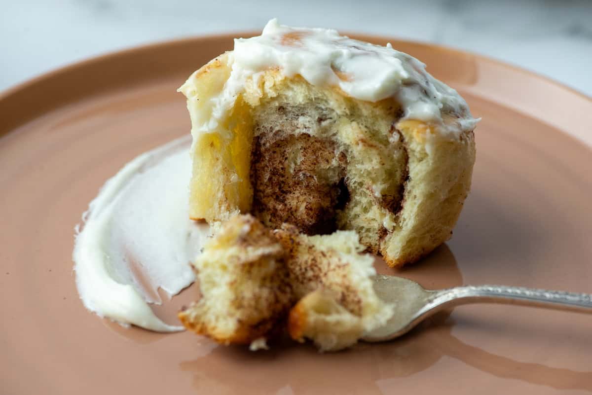 A partially eaten cinnamon roll with white icing on a brown plate, a fork holding a bite-sized piece in front.