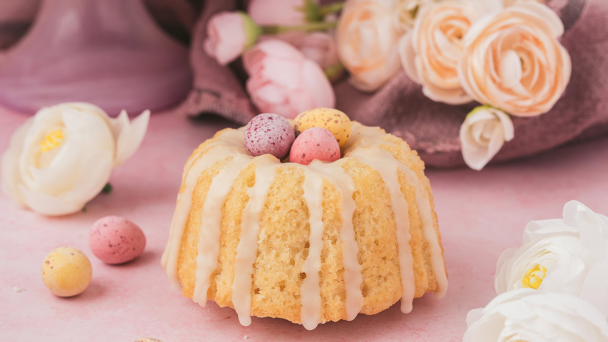 A mini bundt cake with white icing is topped with pastel candy eggs. The cake is surrounded by white and pink flowers on a pink surface.