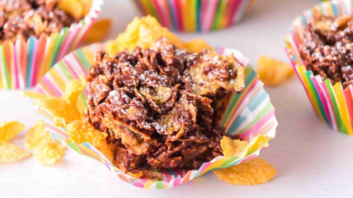 Chocolate cornflake clusters in colorful striped paper cups, topped with a dusting of powdered sugar, with some cornflakes scattered around.