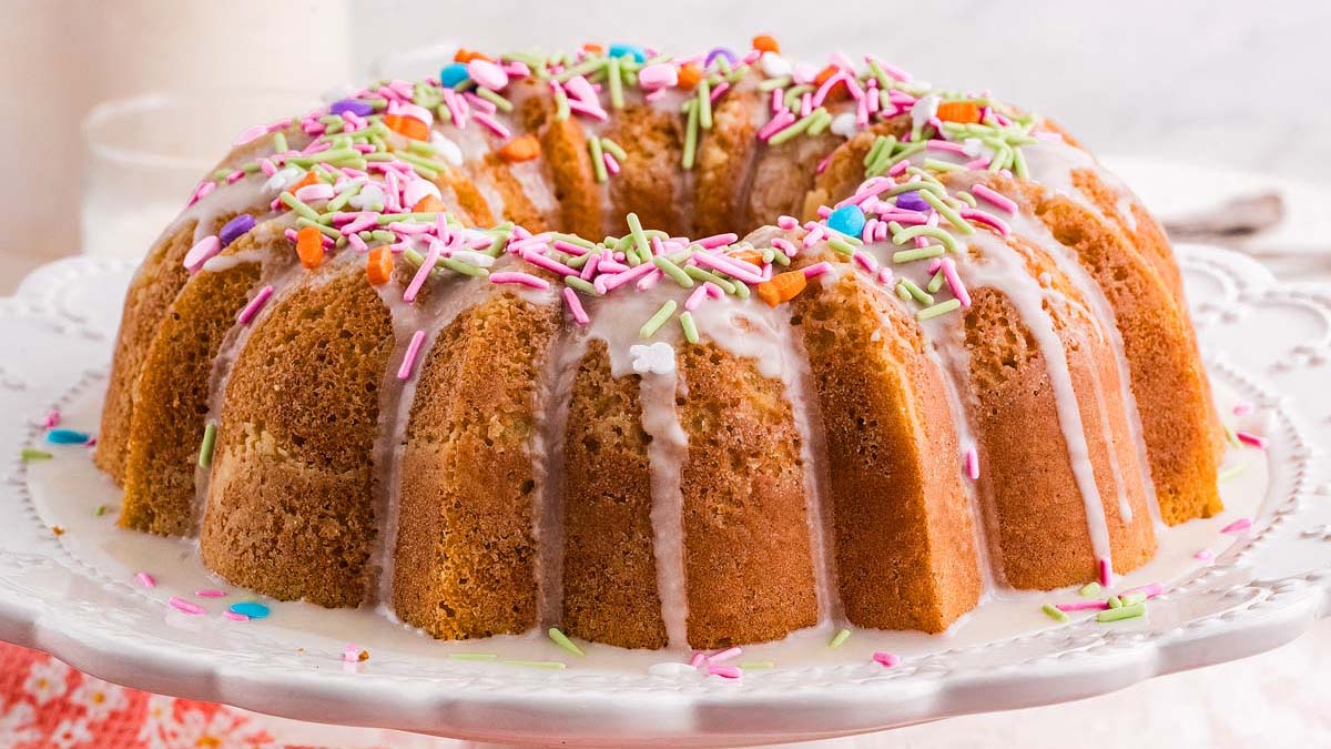 A round bundt cake with white icing drizzled on top and colorful sprinkles, displayed on a white cake stand.