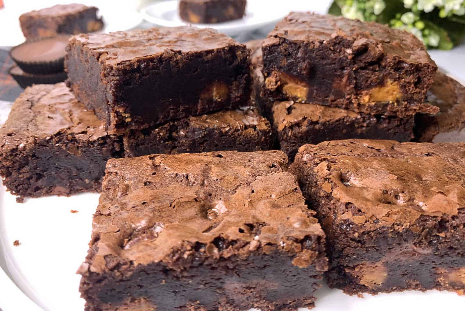 A close-up of several thick, fudgy chocolate brownies stacked on a white plate, with a cracked, shiny top and dense, moist interior.