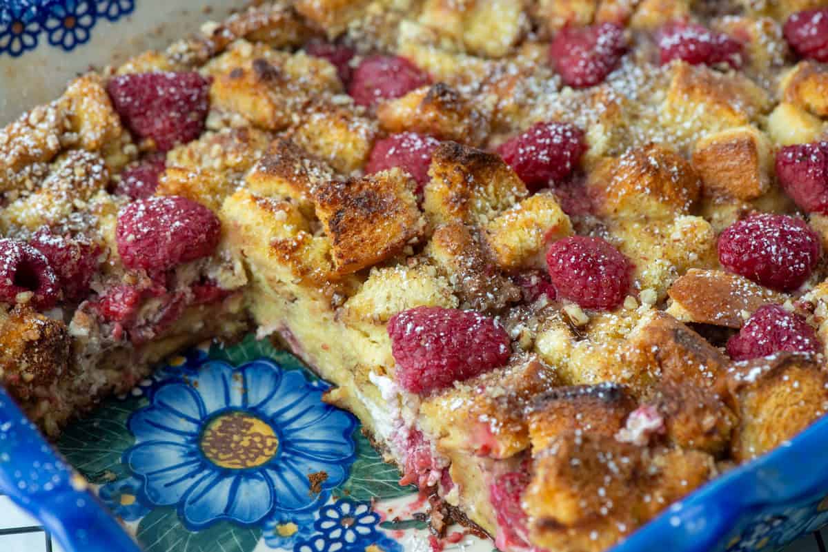 A baked bread pudding with raspberries and powdered sugar, shown in a decorative blue and white baking dish with one serving removed.