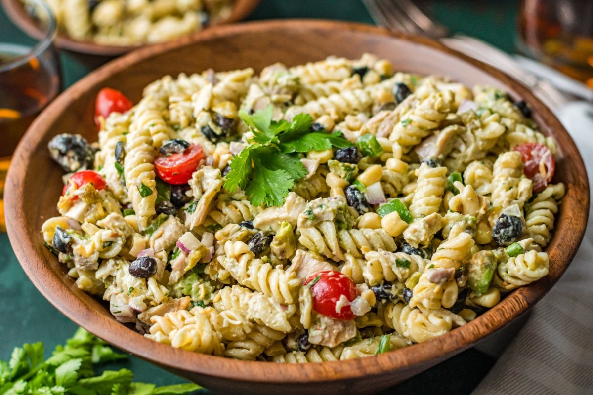 A wooden bowl filled with rotini pasta salad mixed with cherry tomatoes, black olives, corn, herbs, and a creamy dressing, garnished with fresh parsley.