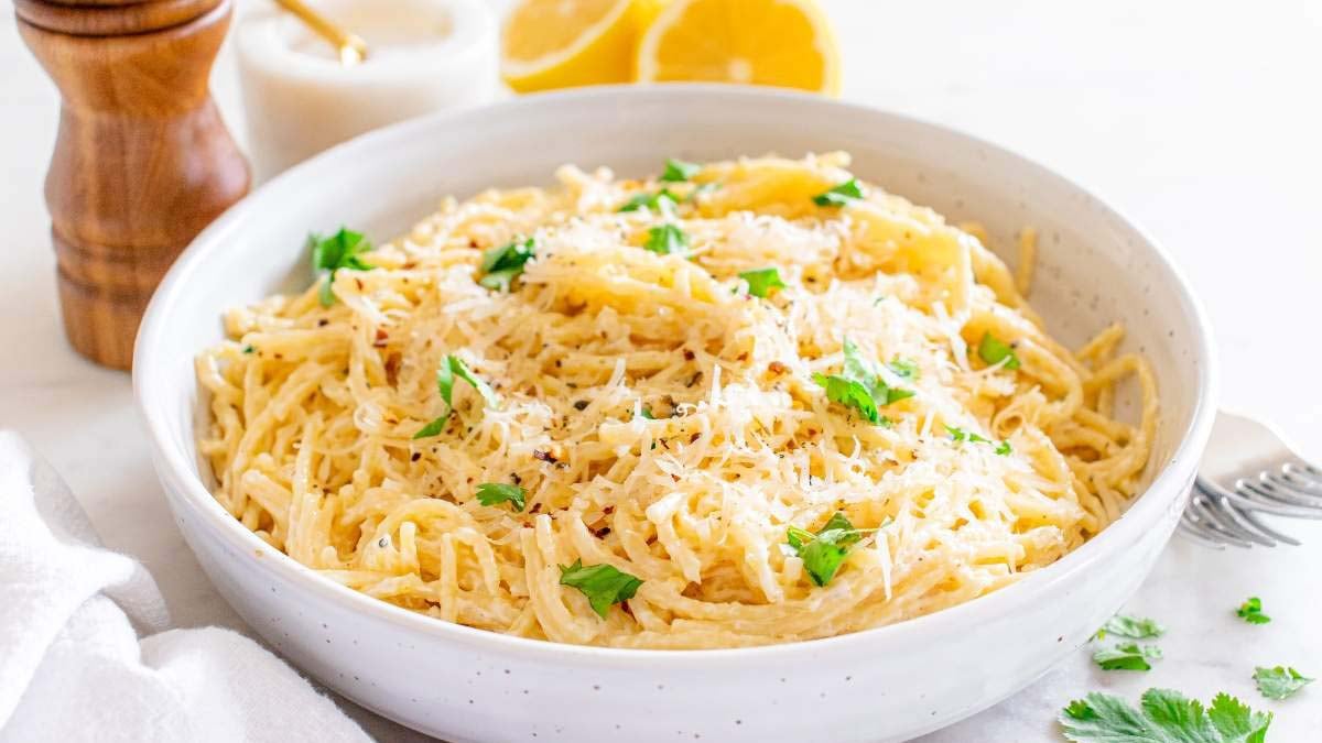 A bowl of creamy spaghetti pasta topped with grated cheese and fresh parsley, with a fork, lemons, and a pepper grinder in the background.