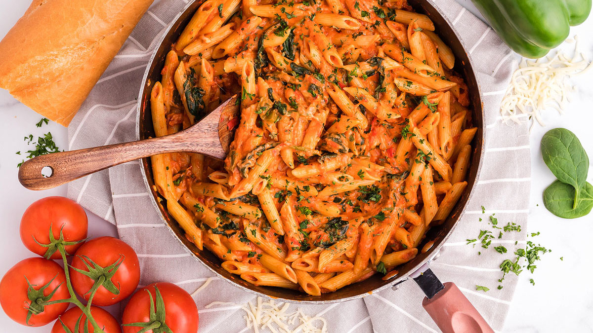 A pan of creamy penne pasta with tomato sauce and herbs sits on a cloth, surrounded by fresh tomatoes, spinach leaves, a baguette, and shredded cheese.