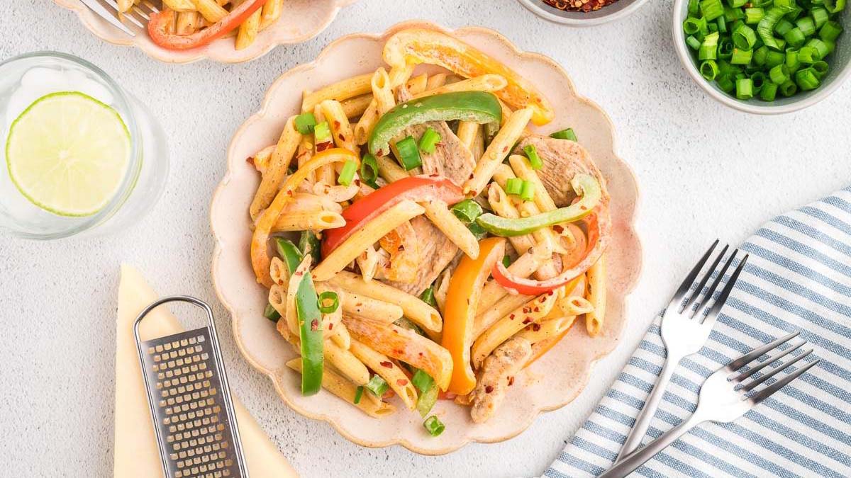 A plate of penne pasta mixed with bell peppers, green onions, and sliced meat, surrounded by small bowls of ingredients, a glass of water with lime, and utensils on a striped napkin.