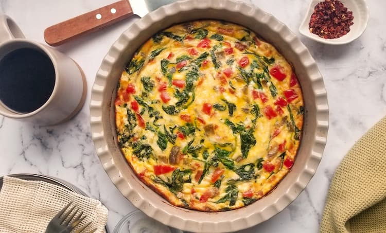 A baked vegetable frittata in a round dish, with spinach, tomatoes, and cheese, next to a cup of coffee, utensils, and a small bowl of red pepper flakes on a marble surface.