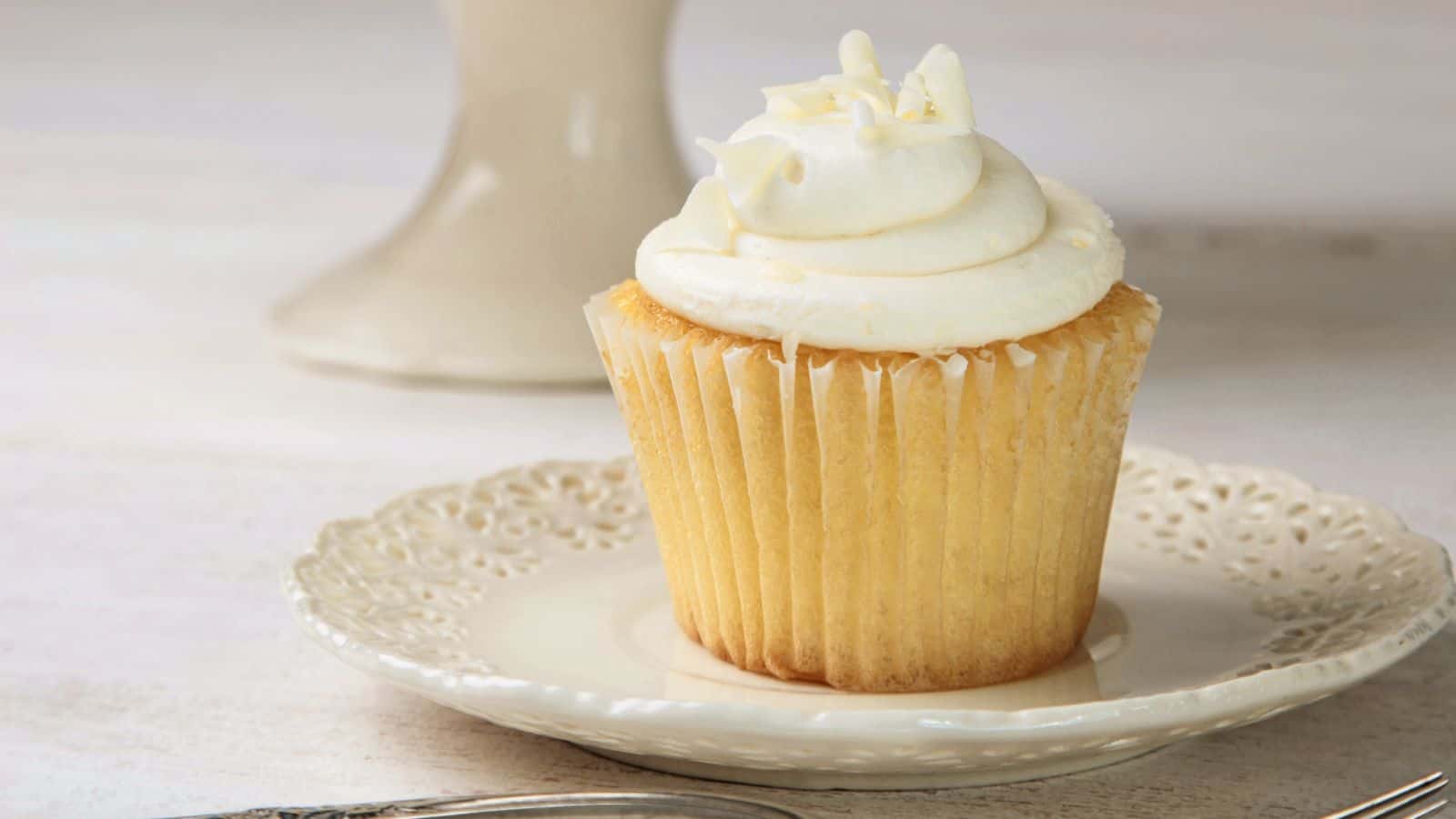 A vanilla cupcake with white frosting sits on a decorative white plate.