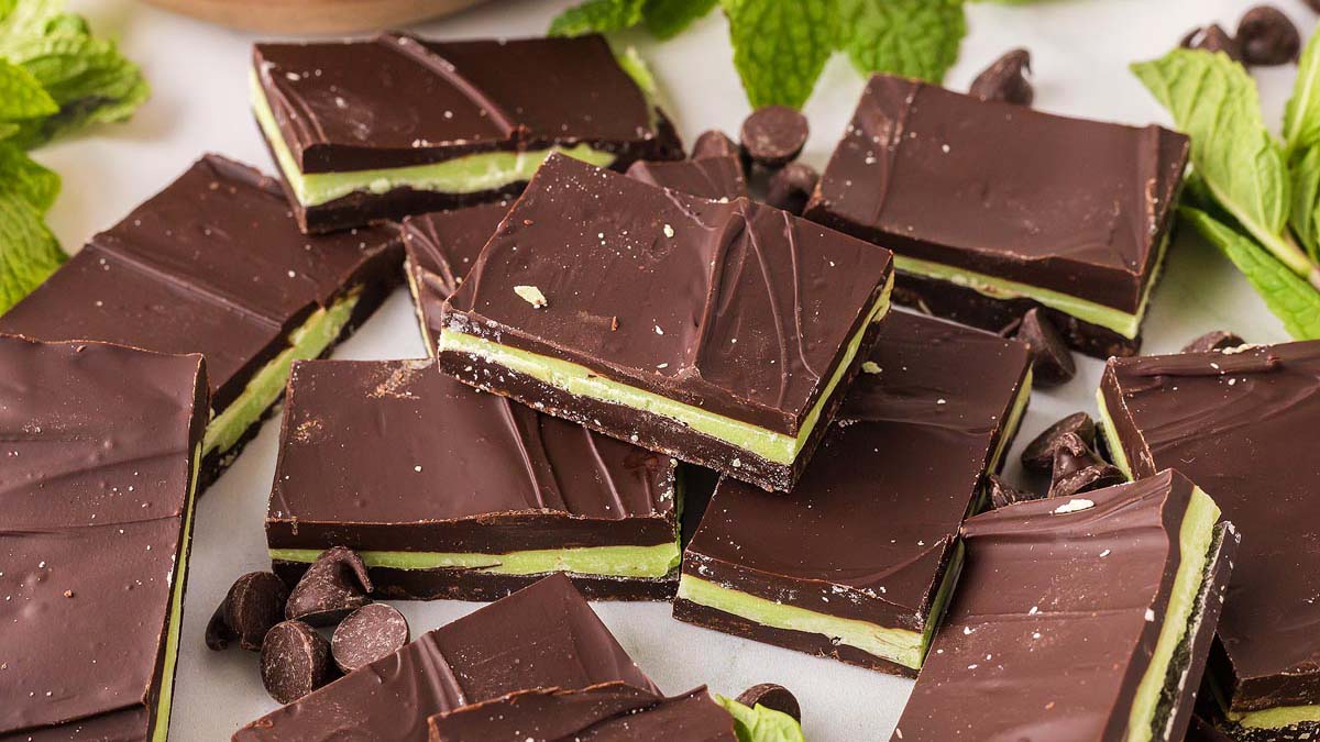 Rectangular pieces of chocolate mint bars with a green mint layer in the center, surrounded by chocolate chips and fresh mint leaves.