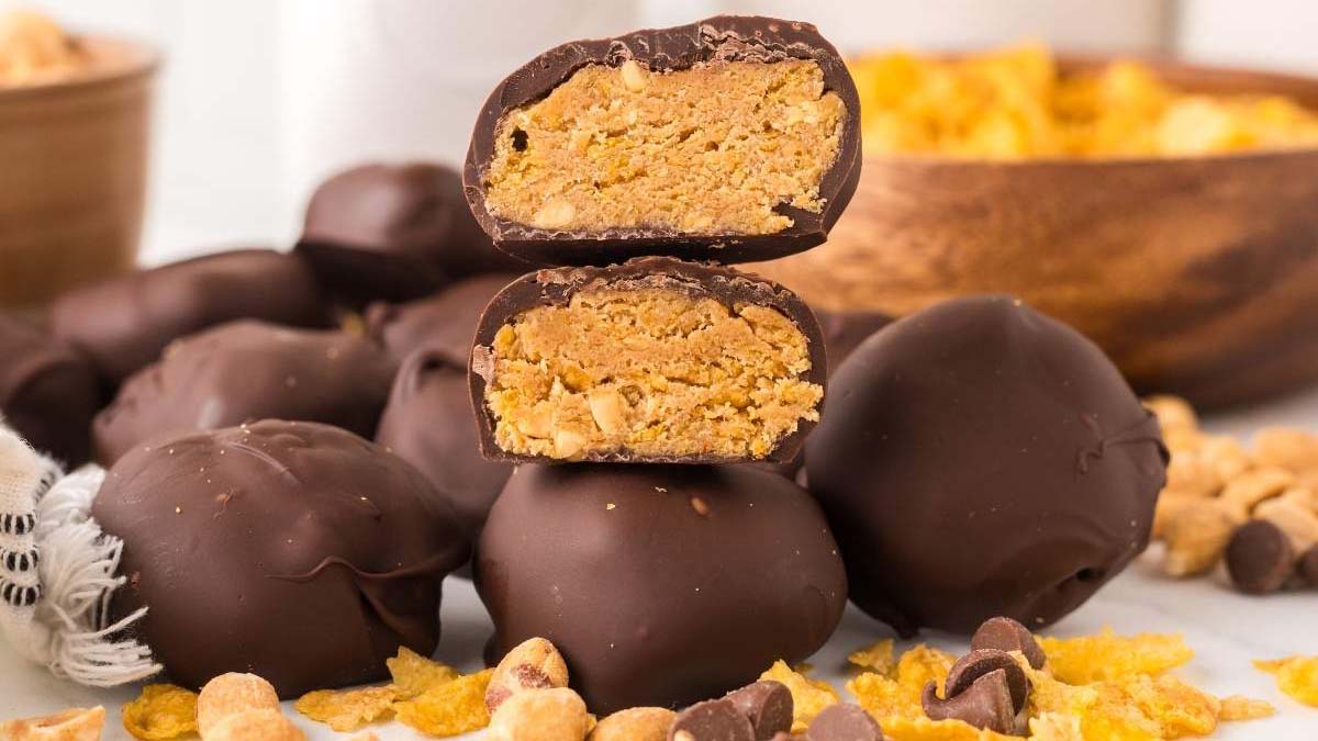 A stack of round chocolate-covered peanut butter balls, with one ball cut in half to show the crumbly peanut butter filling inside. Cornflakes and peanuts are scattered around.