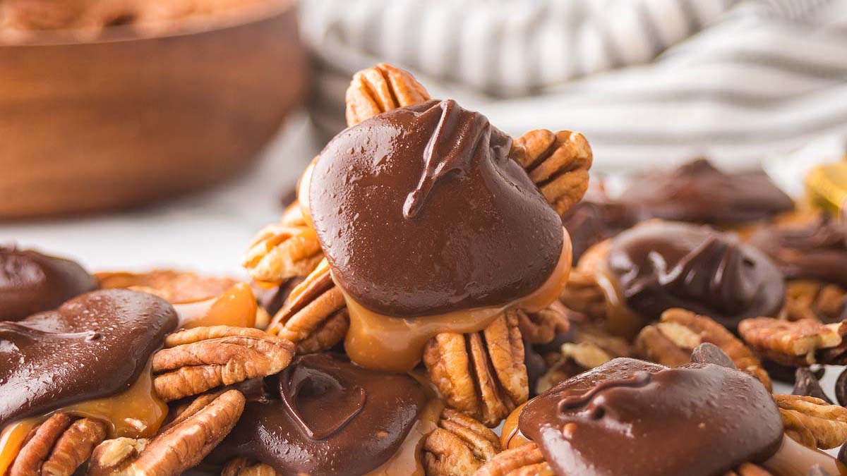 Pecan clusters topped with caramel and coated in chocolate, arranged on a surface with a bowl of pecans in the background.