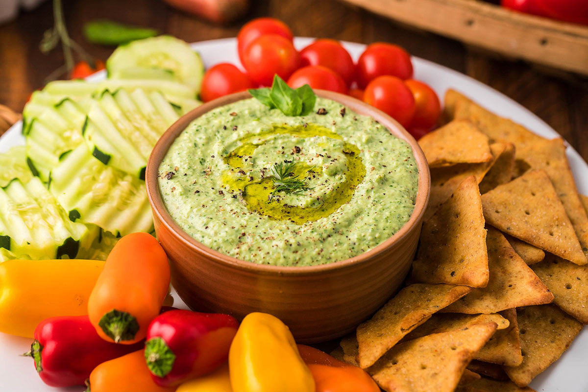 A bowl of green dip is surrounded by sliced cucumbers, cherry tomatoes, mini bell peppers, and triangular crackers on a white plate.
