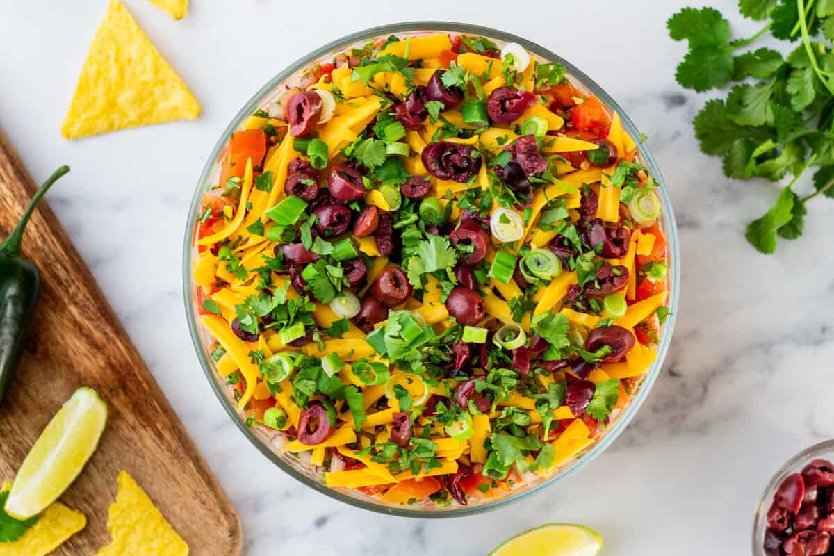 A glass bowl filled with layered dip topped with shredded cheese, chopped olives, green onions, and cilantro, surrounded by tortilla chips, lime wedges, and a jalapeรฑo.