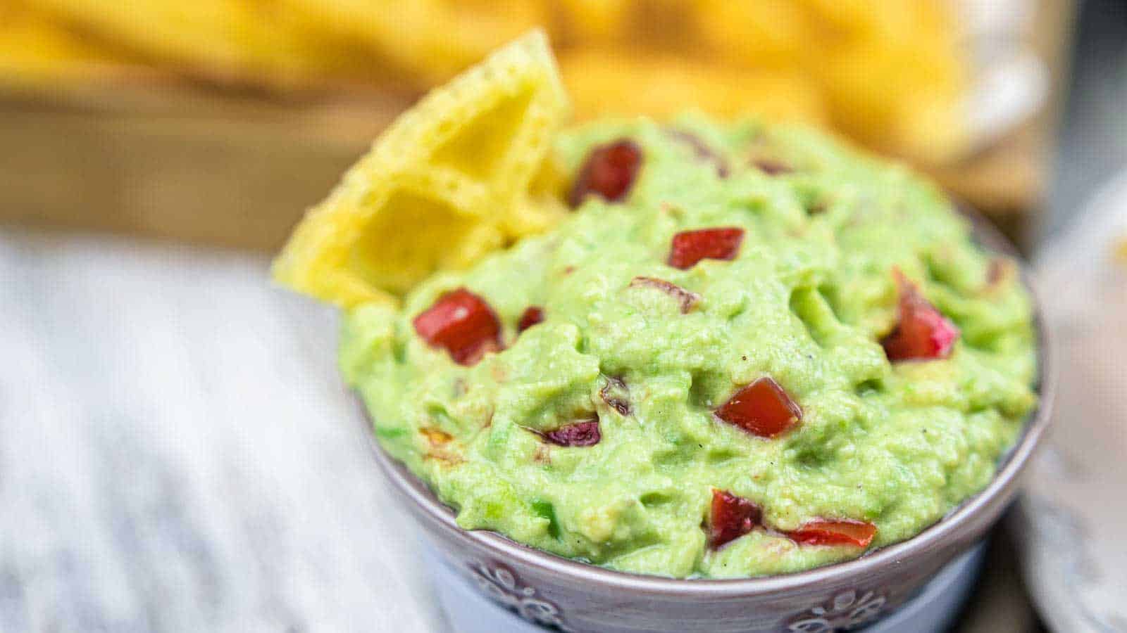 A bowl of guacamole with diced tomatoes, garnished with a tortilla chip on the side.