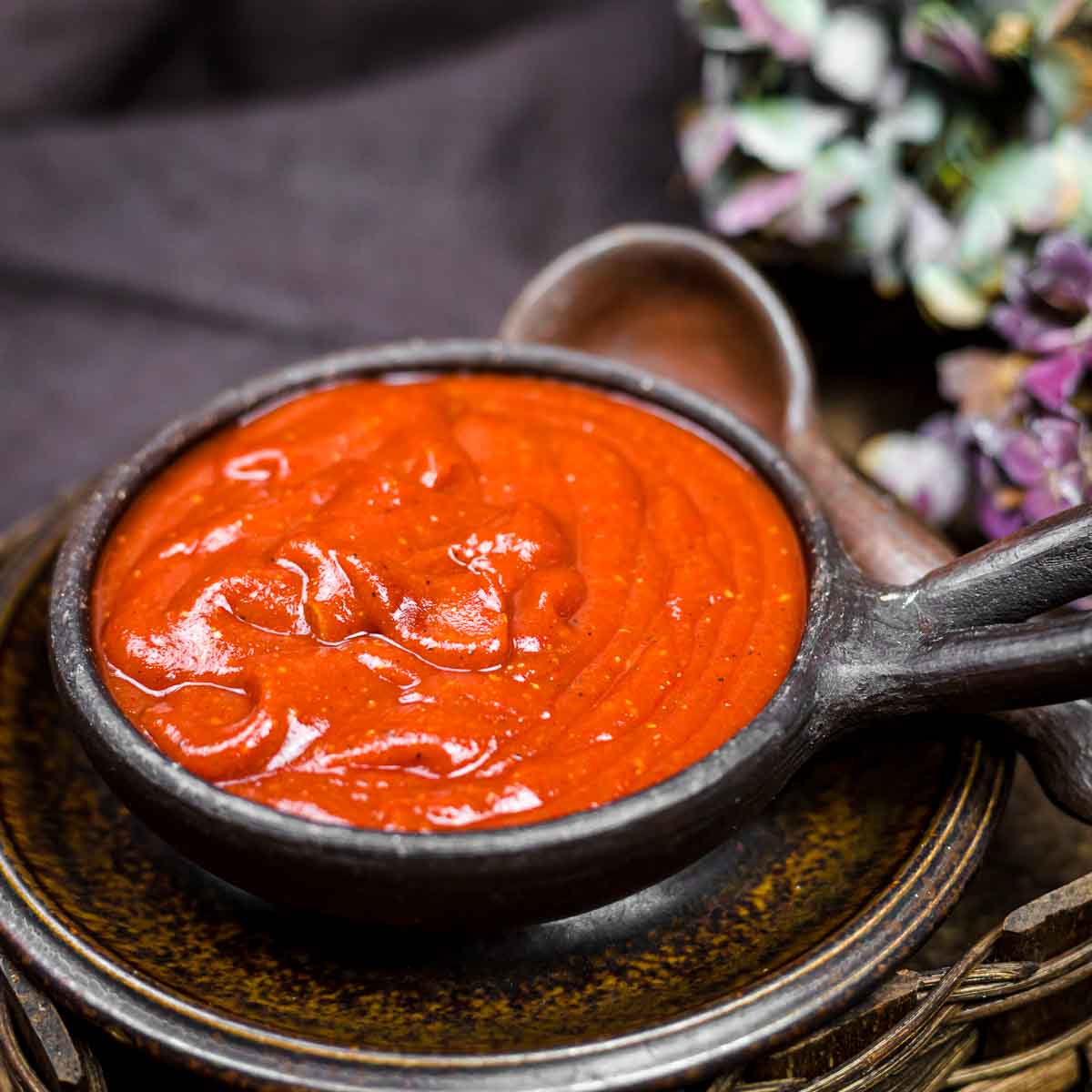 A small dark bowl filled with thick, red sauce sits on a matching plate with a wooden spoon beside it; flowers are visible in the background.