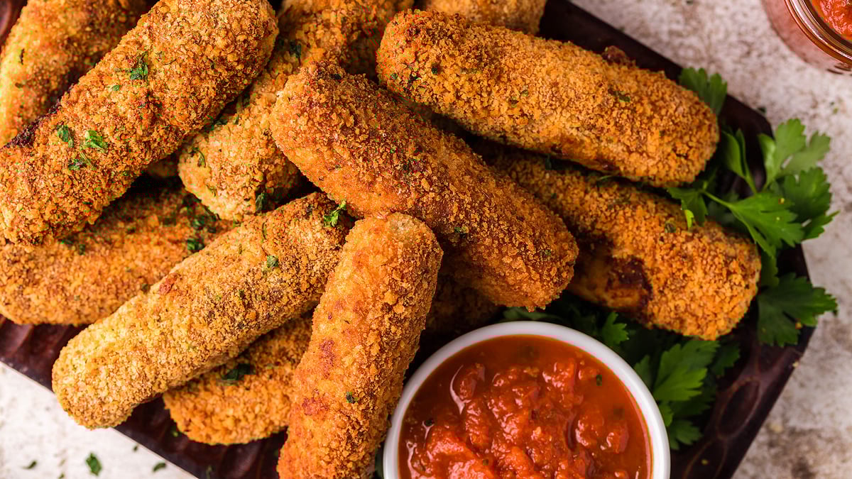 A pile of breaded and fried mozzarella sticks on a wooden platter, served with a bowl of marinara sauce and garnished with fresh parsley.