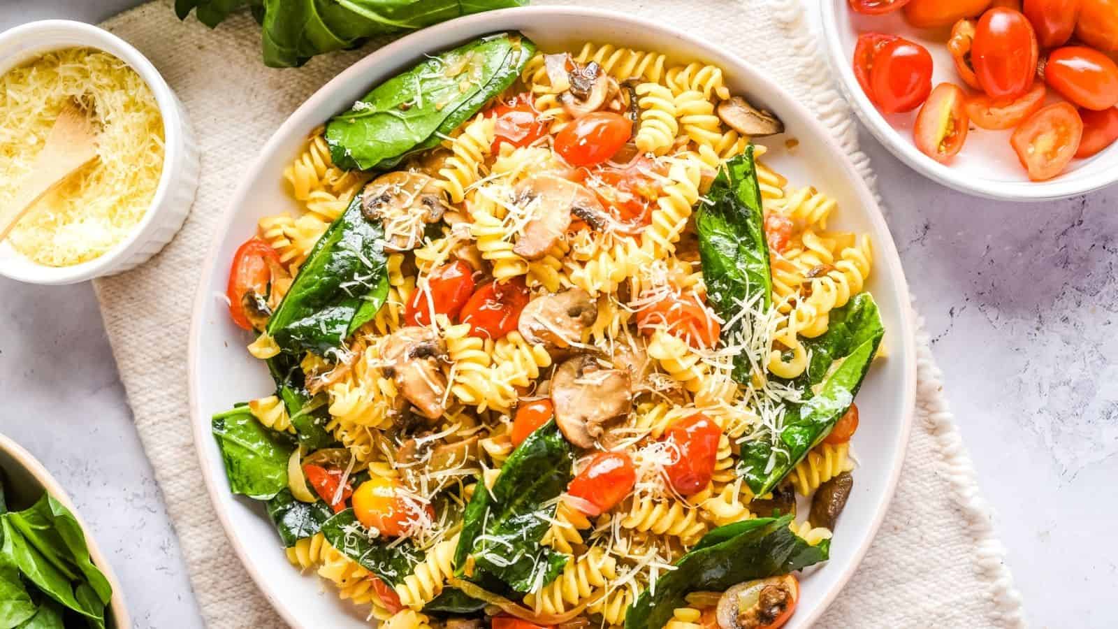 A plate of rotini pasta with spinach, cherry tomatoes, mushrooms, and grated cheese, surrounded by bowls of grated cheese, fresh spinach, and cherry tomatoes.