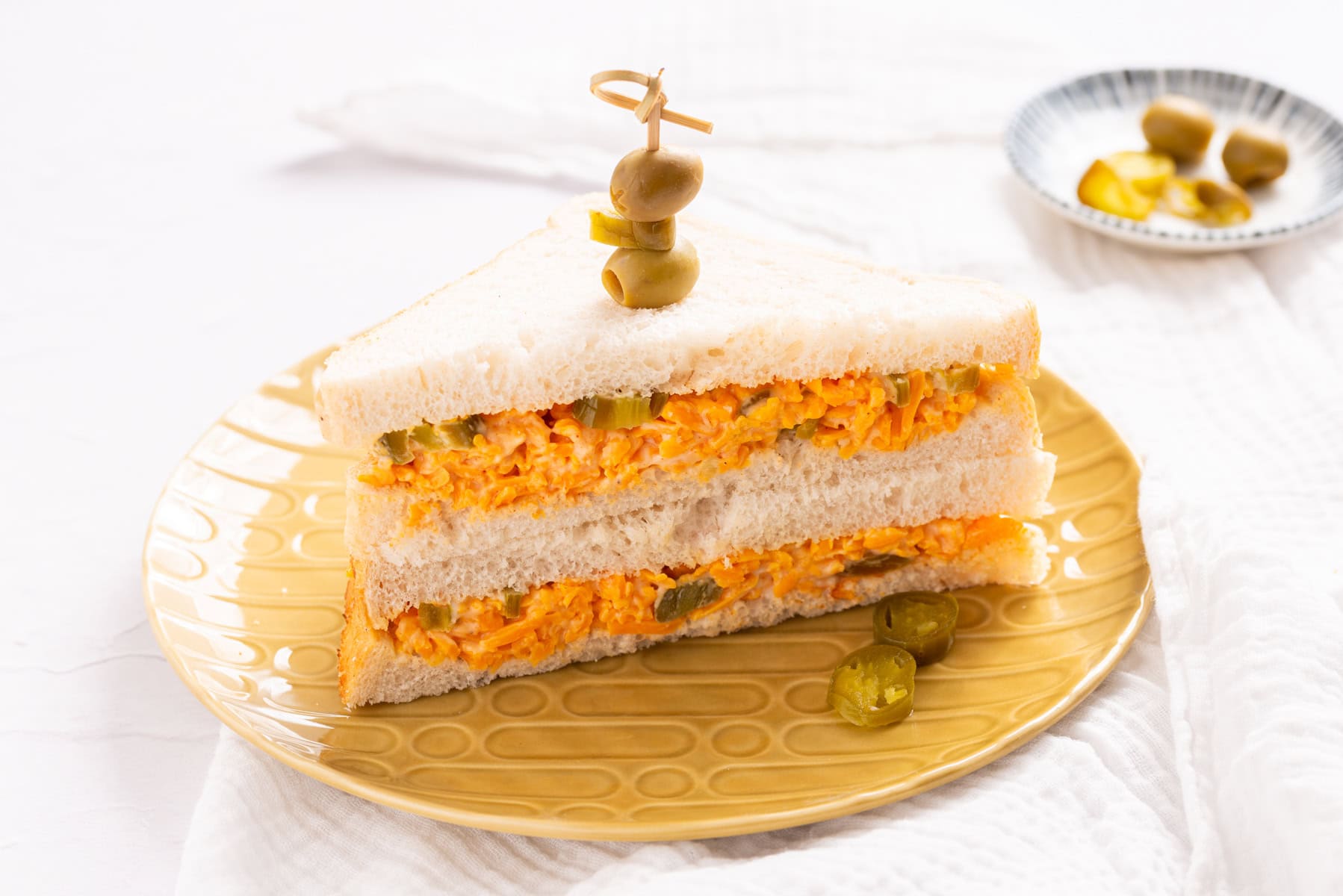A stacked sandwich with shredded filling and pickles, garnished with green olives on a toothpick, served on a yellow plate.