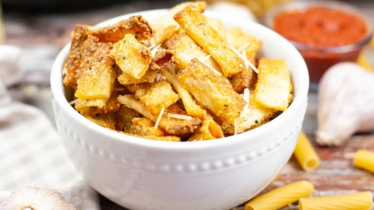 A white bowl filled with crispy baked pasta pieces, garnished with grated cheese and herbs, with a dish of red sauce and garlic in the background.