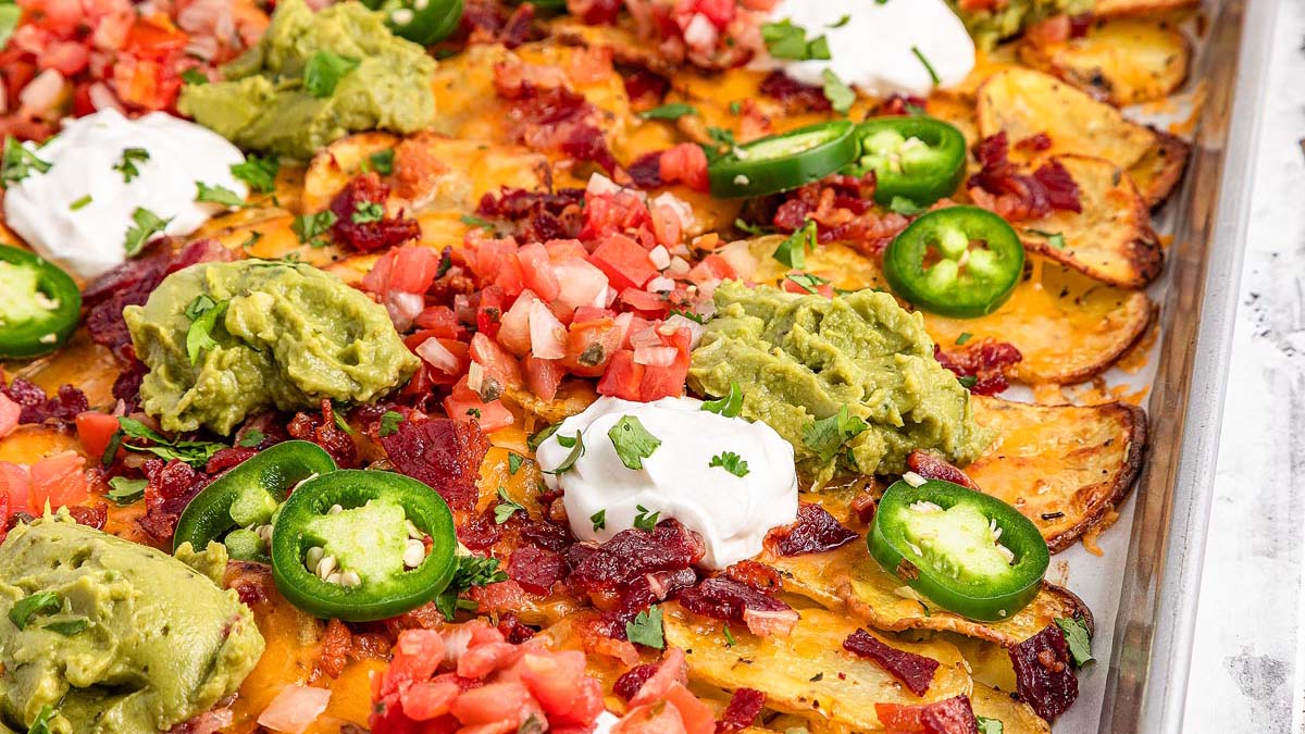 Sheet pan loaded nachos topped with melted cheese, sour cream, guacamole, diced tomatoes, bacon bits, jalapeรฑo slices, and chopped herbs.