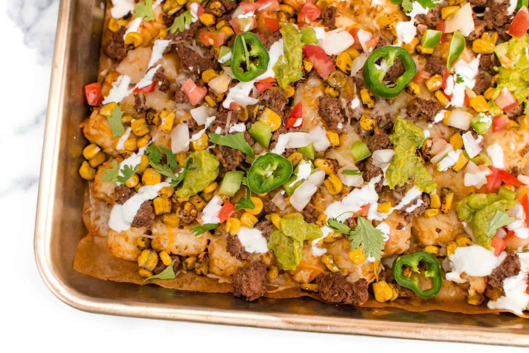 A baking tray filled with enchiladas topped with ground beef, corn, jalapeรฑo slices, guacamole, diced onions, tomatoes, cilantro, and drizzled with white sauce.