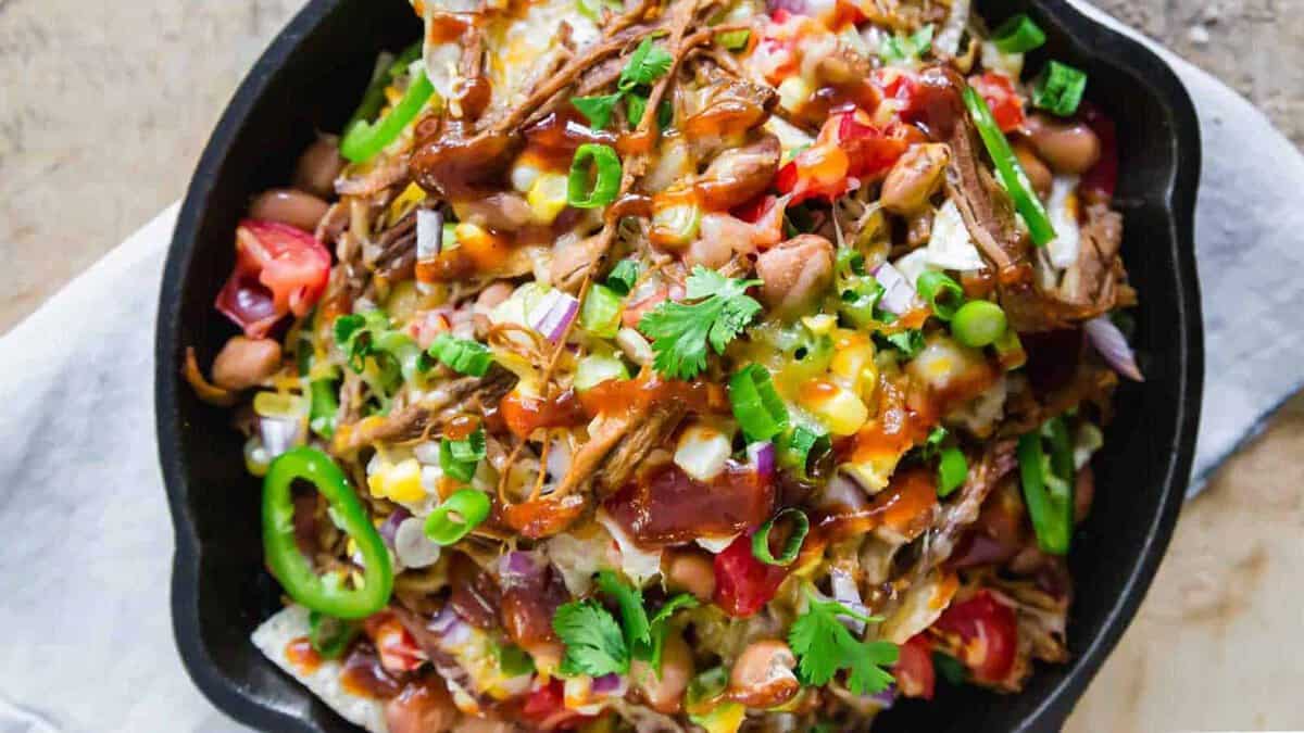 A cast iron skillet filled with loaded nachos topped with shredded meat, beans, cheese, jalapeรฑos, onions, tomatoes, green onions, and cilantro.