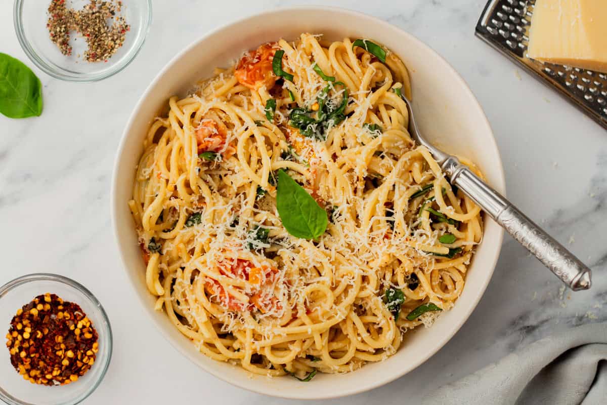 A bowl of spaghetti topped with grated cheese, cherry tomatoes, spinach, and a basil leaf, with a fork in the bowl and small dishes of spices on the side.