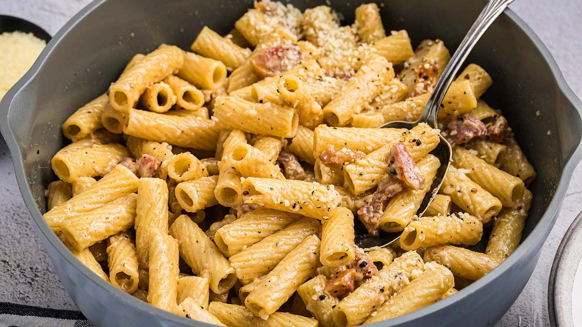 A pan filled with cooked rigatoni pasta mixed with a creamy sauce, bacon pieces, grated cheese, and black pepper, with a spoon resting inside.